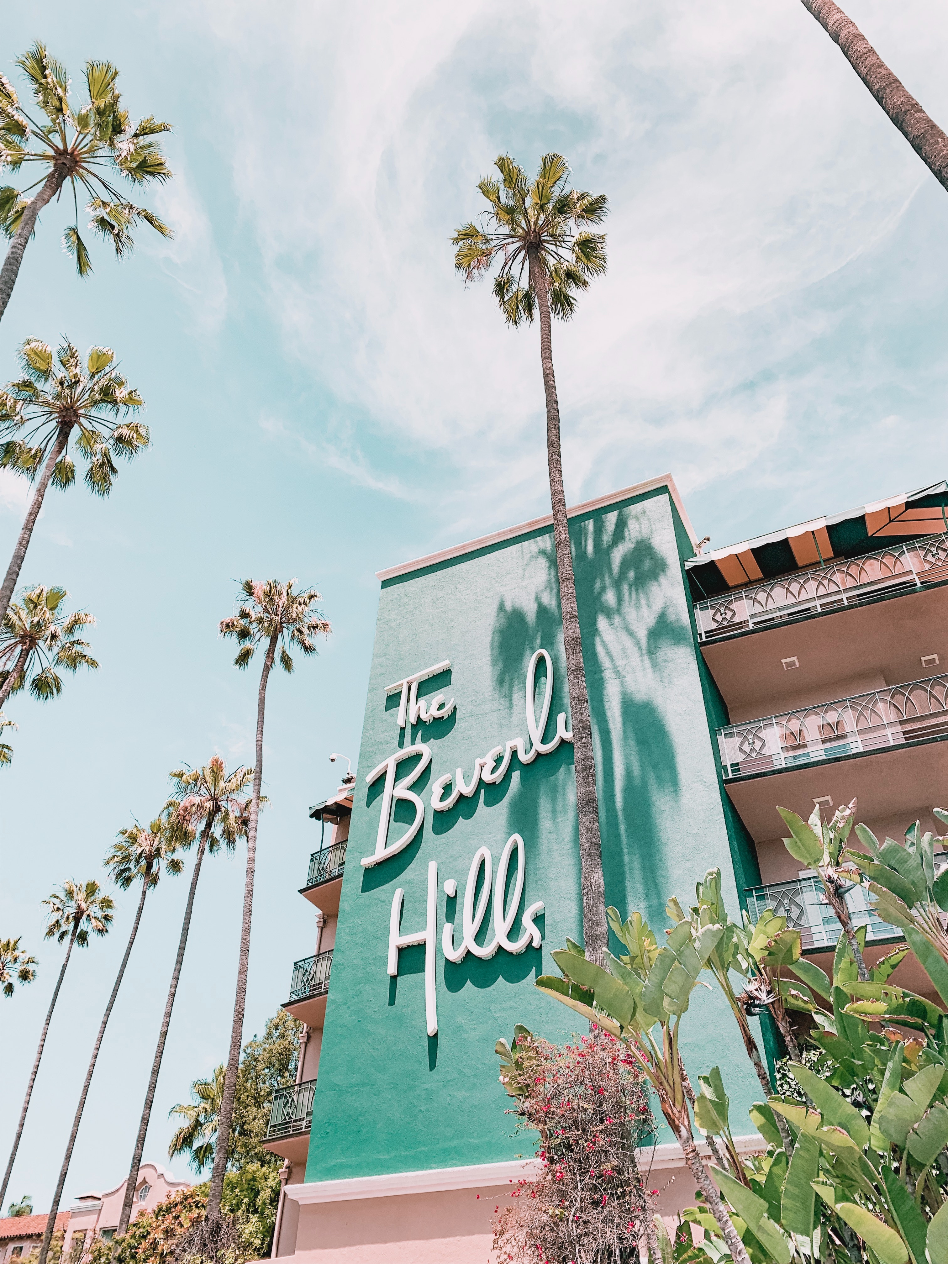 A photo showing the teal exterior of the Beverly Hills hotel against a blue sky, surrounded by palm trees. 