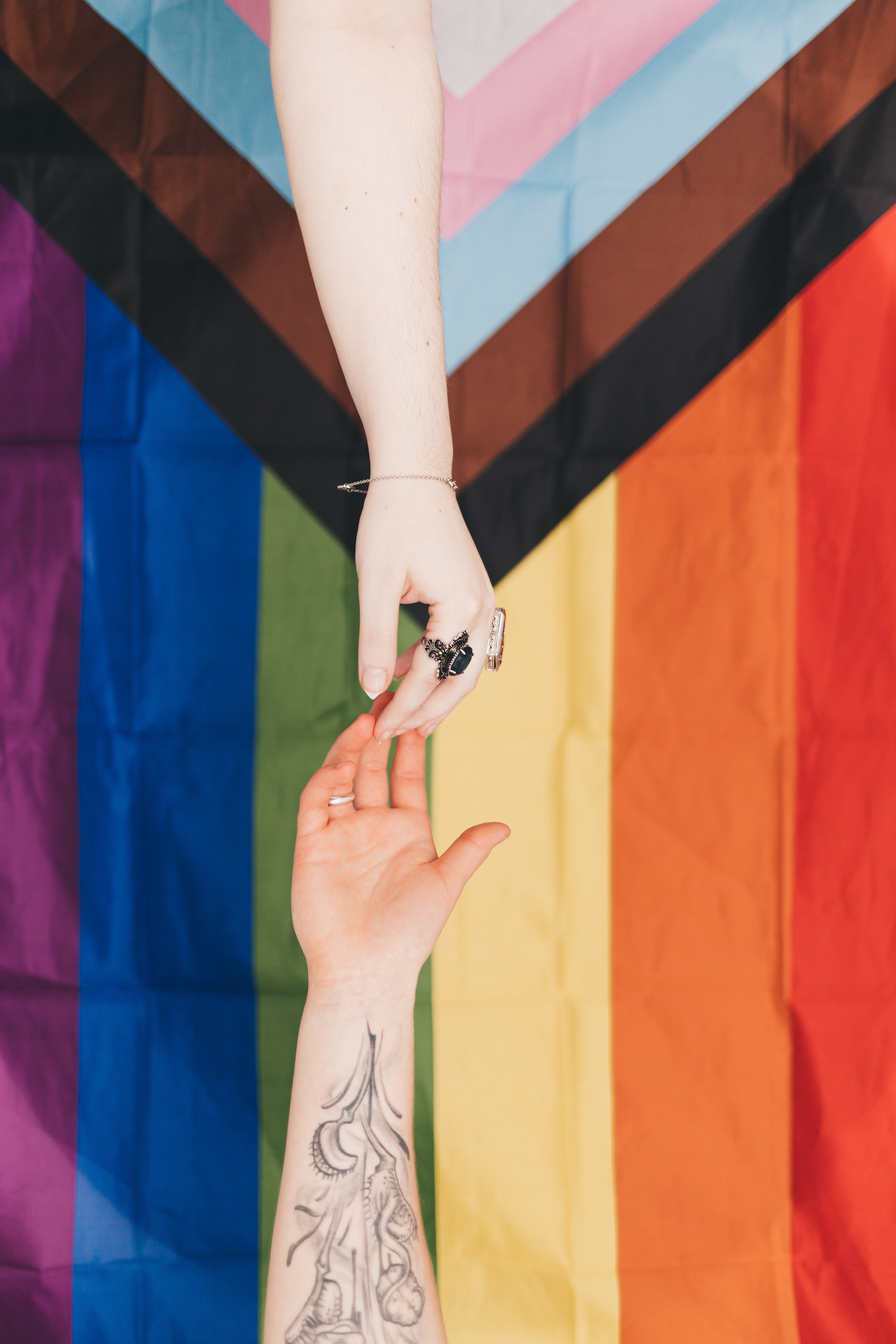 two white hands reaching for one another over the Progress Pride LGBTQ+ flag 