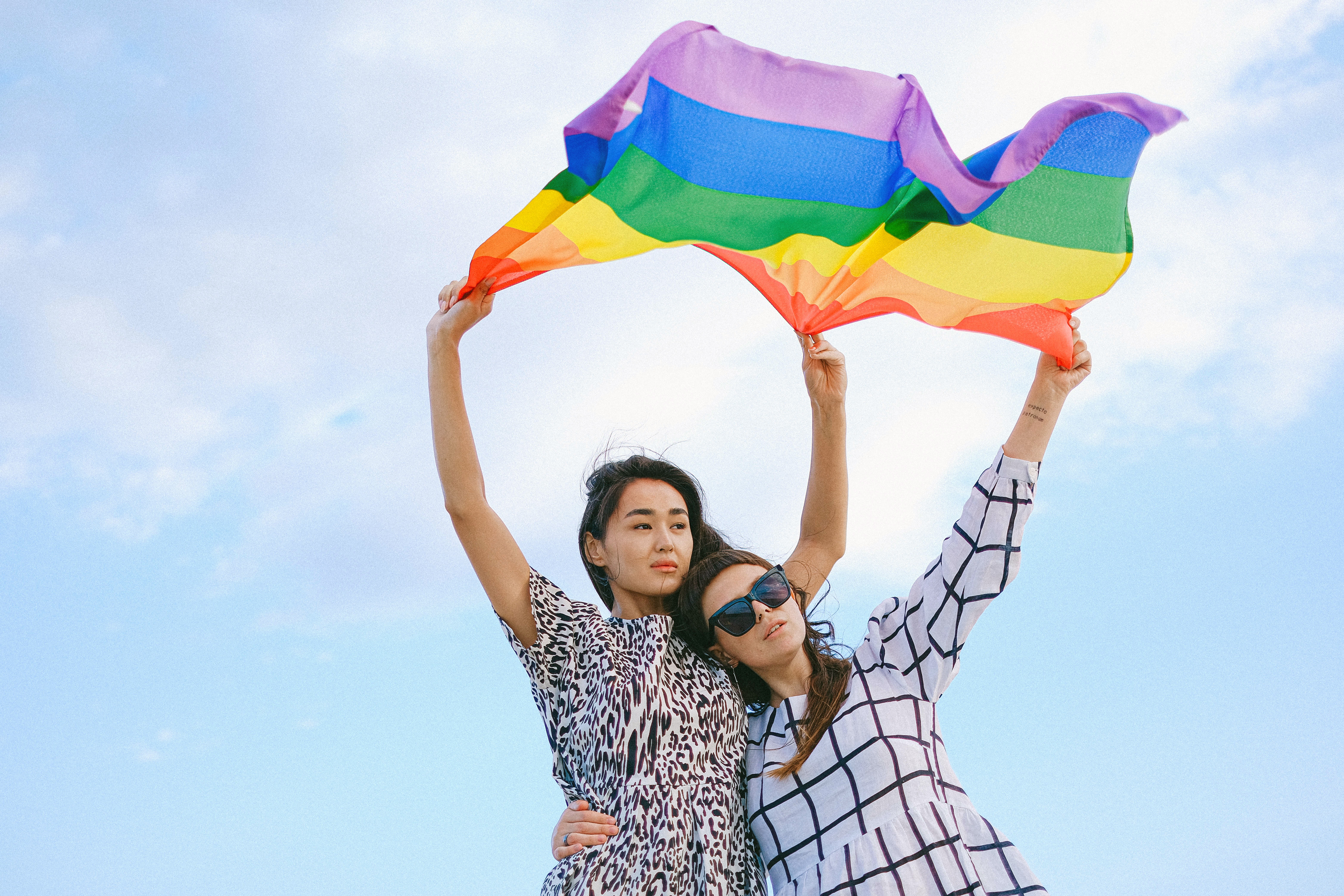 Two queer Asian American women holding a pride flag overhead, with a blue sky in the background. One has her arm around the other's waist.