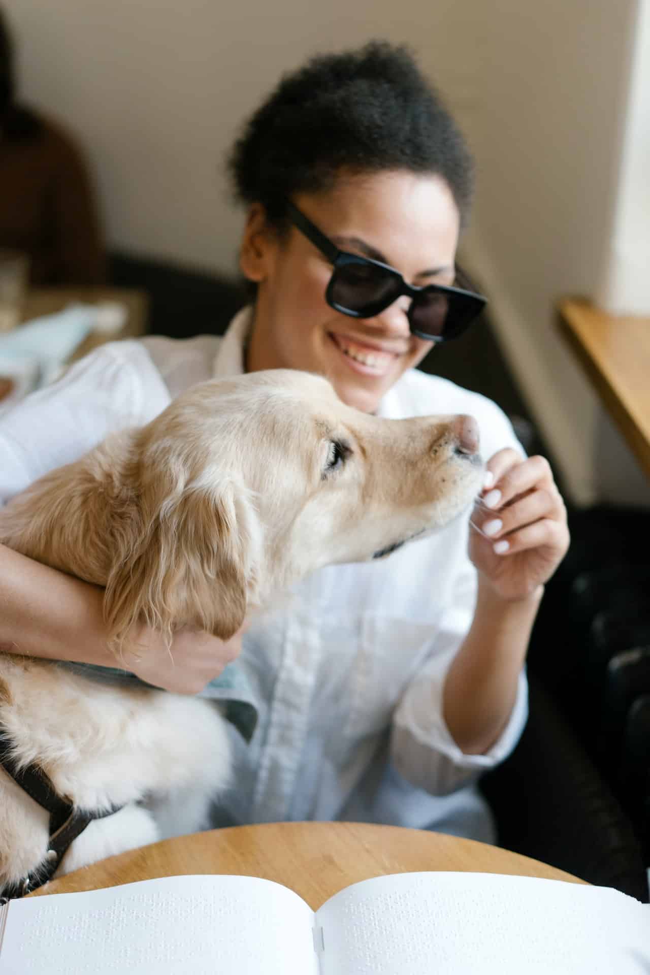 A blind Black woman wearing dark glasses sits at a table with a braille book open in front of her. She is smiling and feeding a treat to her service animal, a golden retriever.