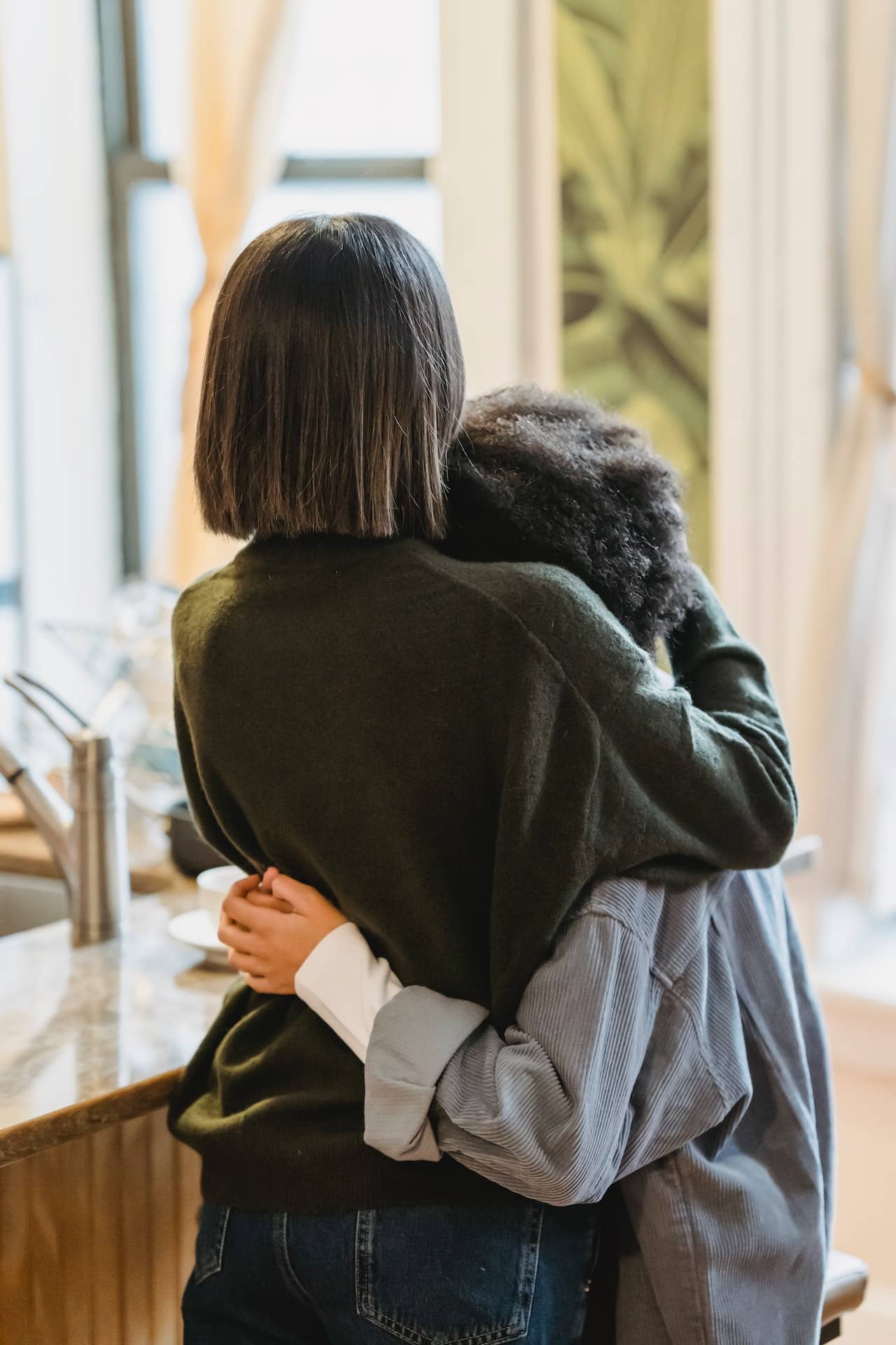 Stock photo of two people hugging from behind, one with a short straight bob, one with an afro hairstyle. They appear to be supporting each other.