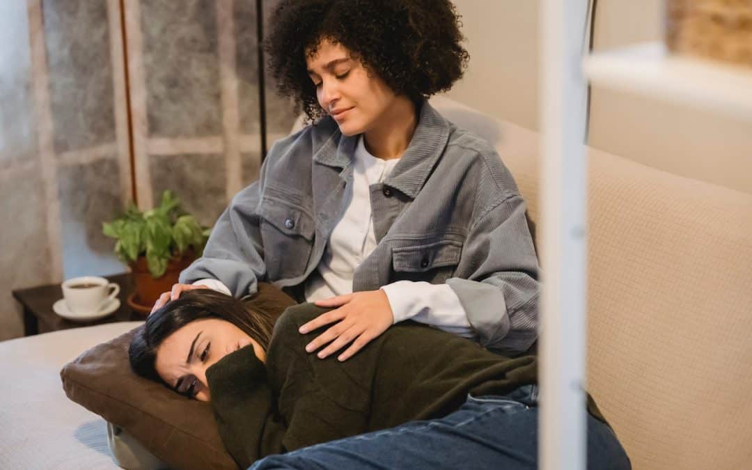 Stock photo of a woman providing support to another grieving woman, who is laying with her head on a pillow on the first woman's lap.