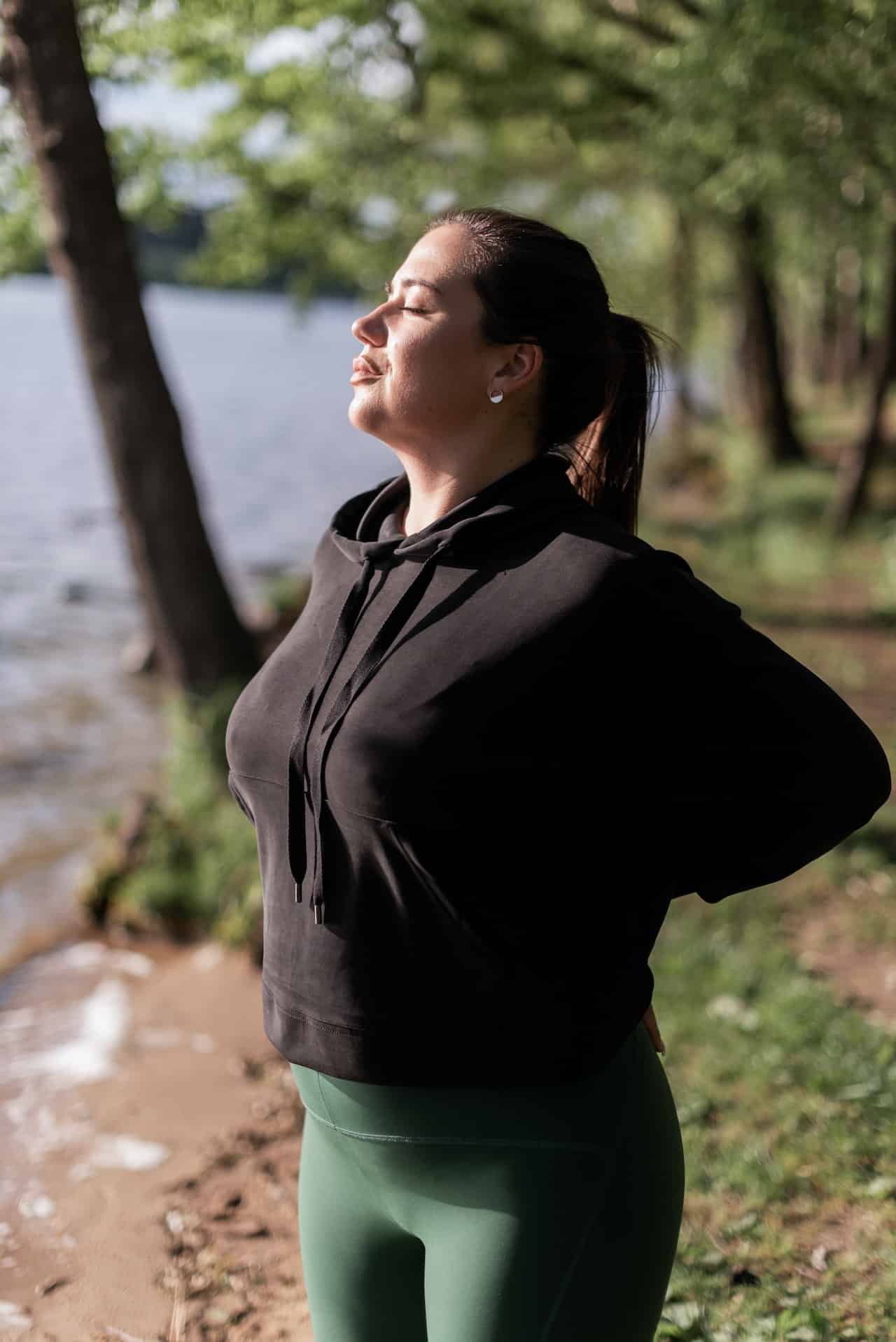 A woman in casual workout attire takes a deep breath, eyes closed. She is standing outdoors with a lake and trees visible in the background.