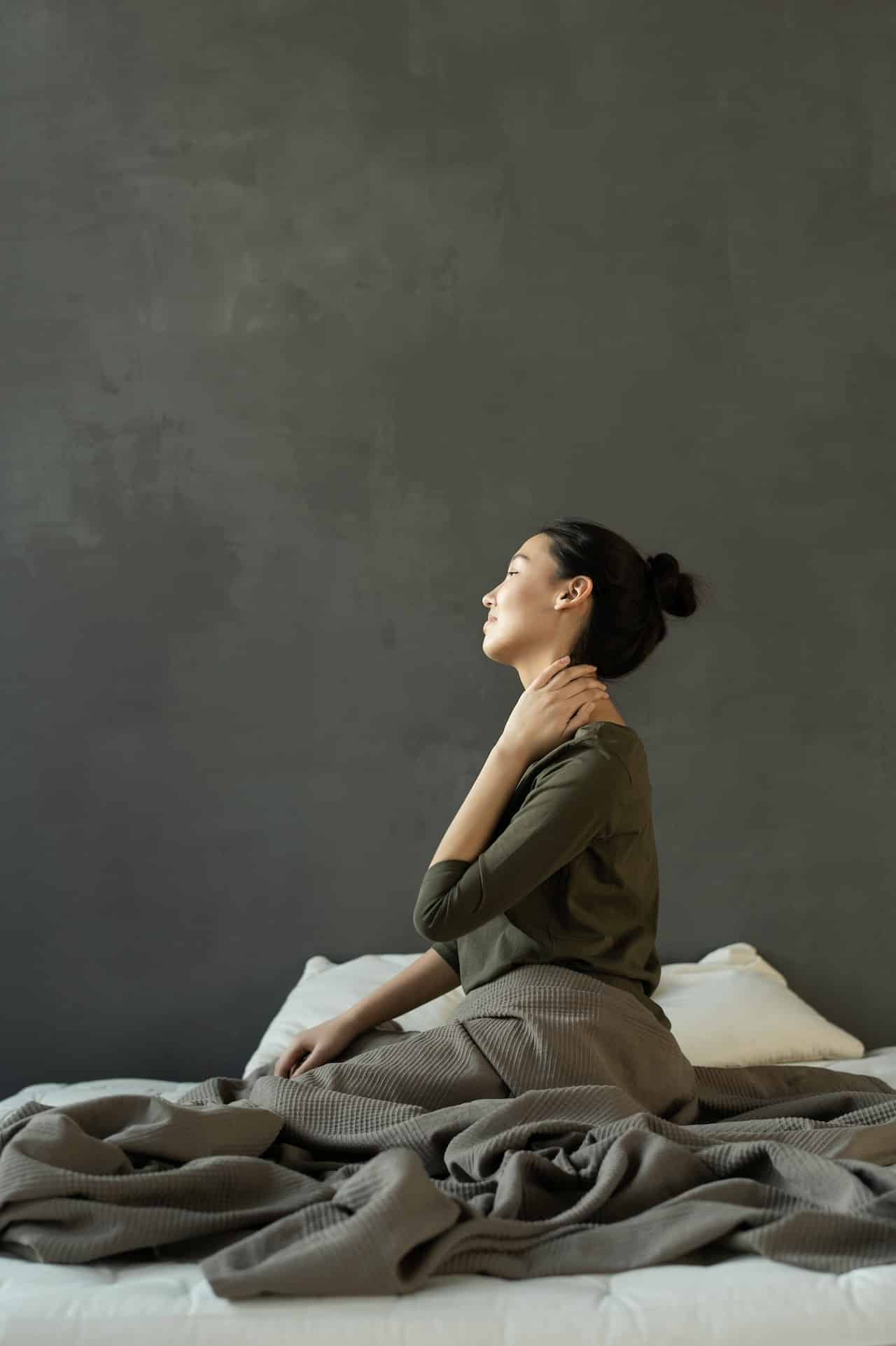 An asian american woman sits up in bed. Turning away from the camera, she is stretching her neck as though to relieve muscle tension.