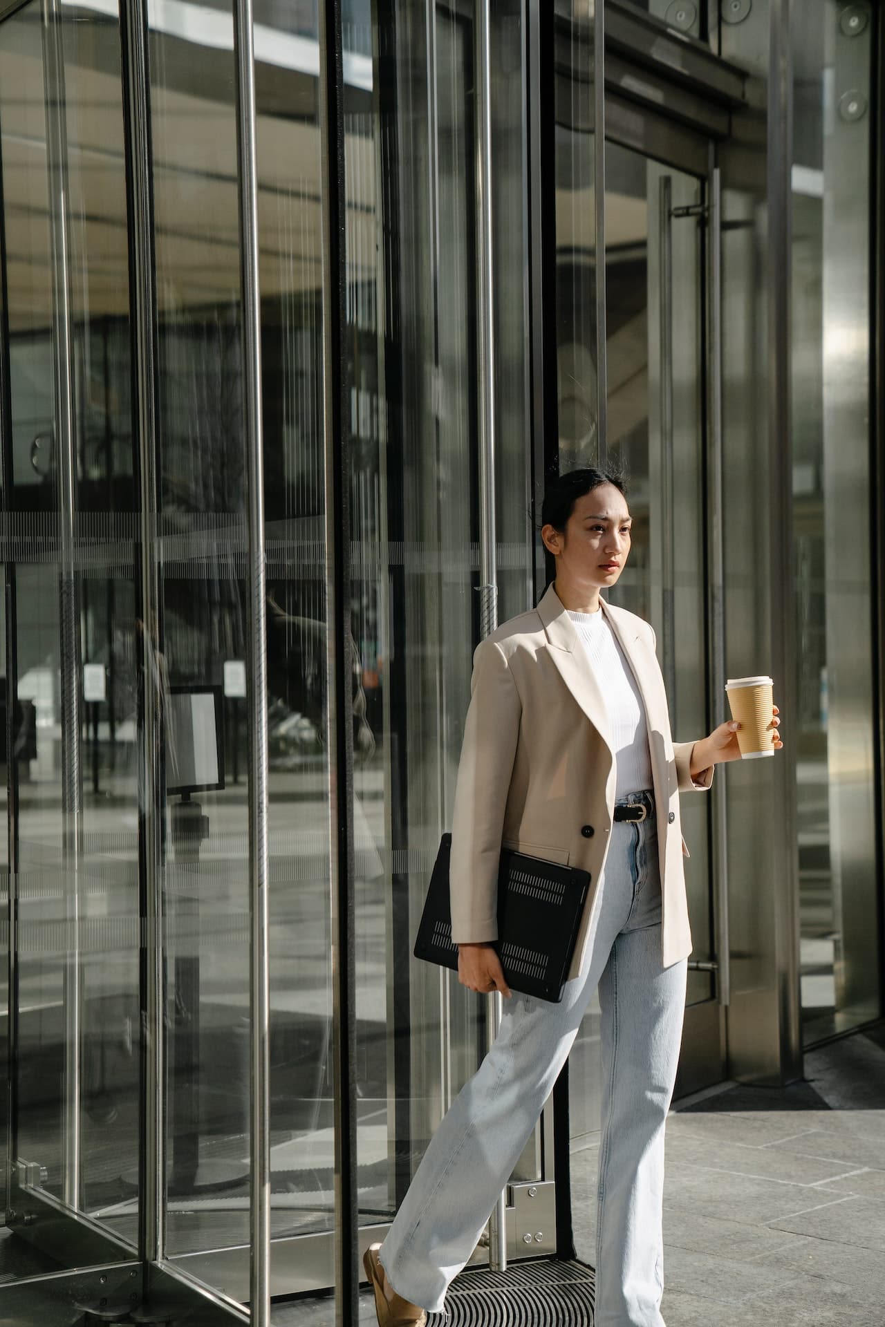 A confident Asian woman in business attire walks out the door of a corporate office with a cup of coffee.