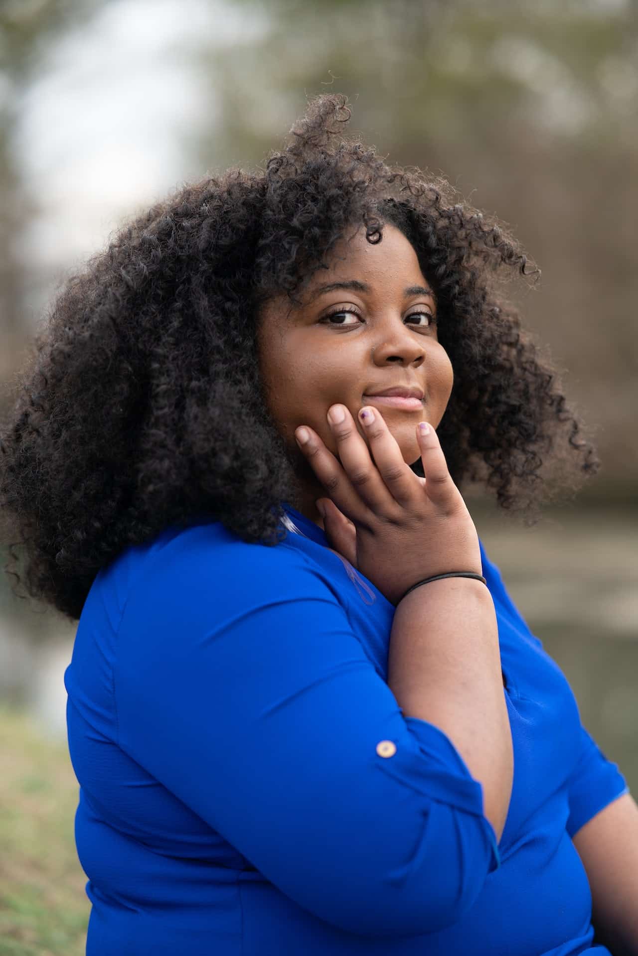 A Black woman with curly hair looks at the camera with her hand on her face. She is wearing a blue shirt.