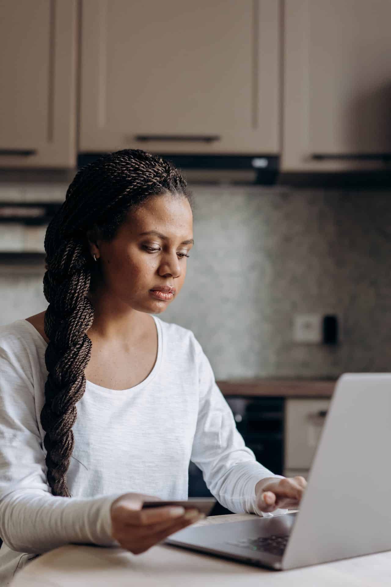 Black woman with braids pulled back sits at a laptop, with a card in her hand, as though she is checking her insurance benefits on her computer.