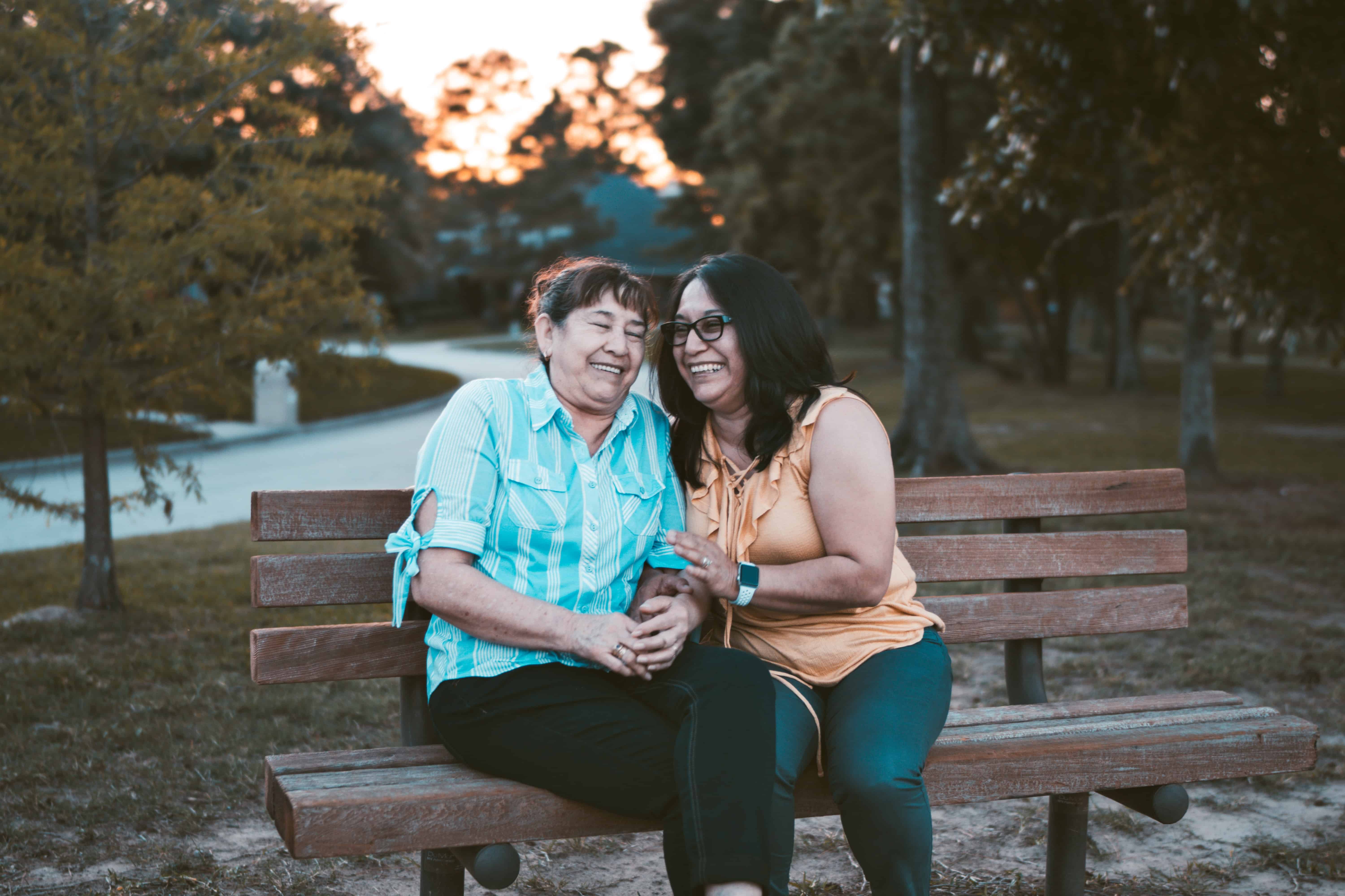 Two Latina women, one older and one middle aged, sitting and laughing on a park bench as the sun sets.
