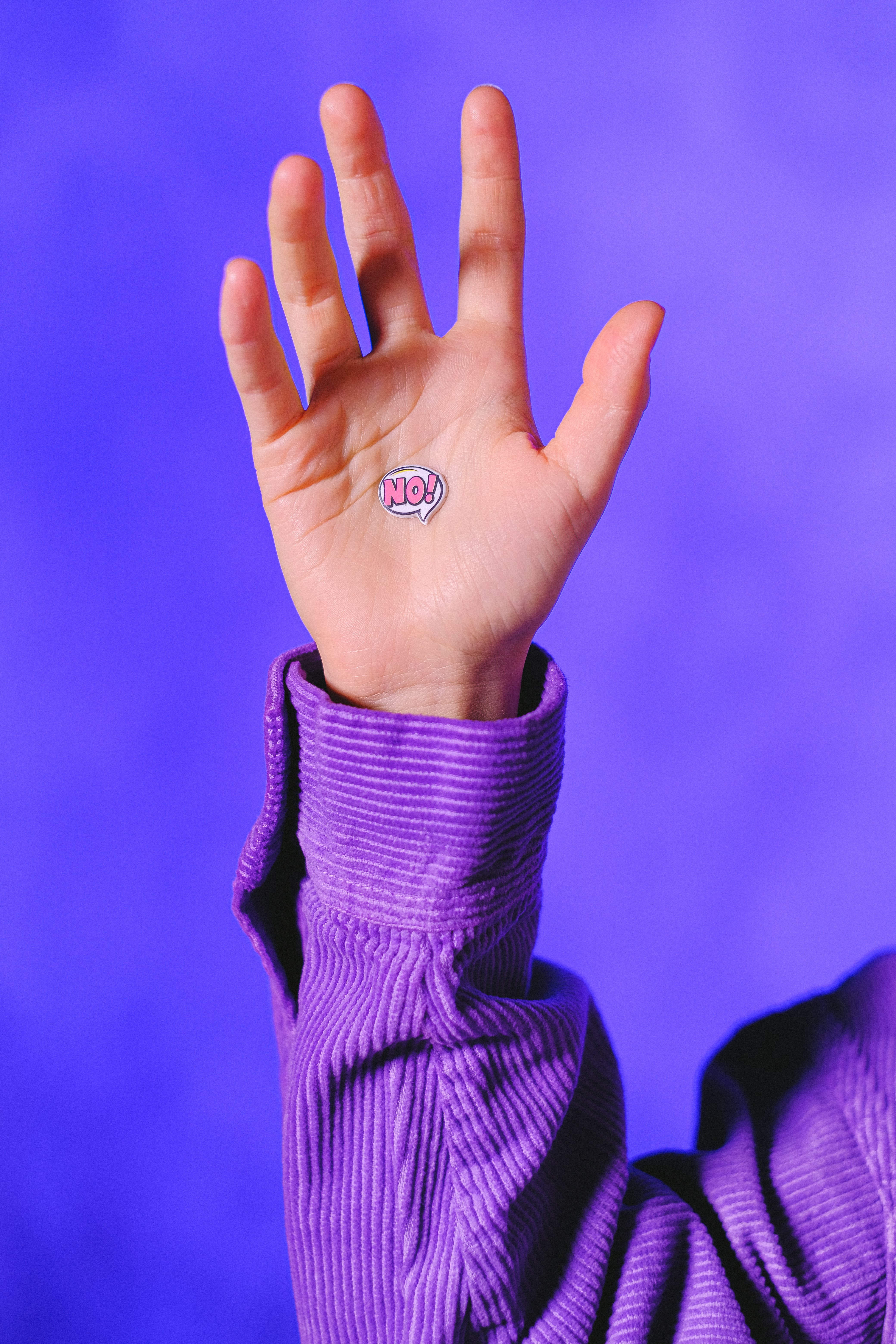 A close up of the raised hand of a person wearing a long sleeved purple shirt, in front of a purple wall, with a small sticker on their palm that reads "No!" in a comic book font.