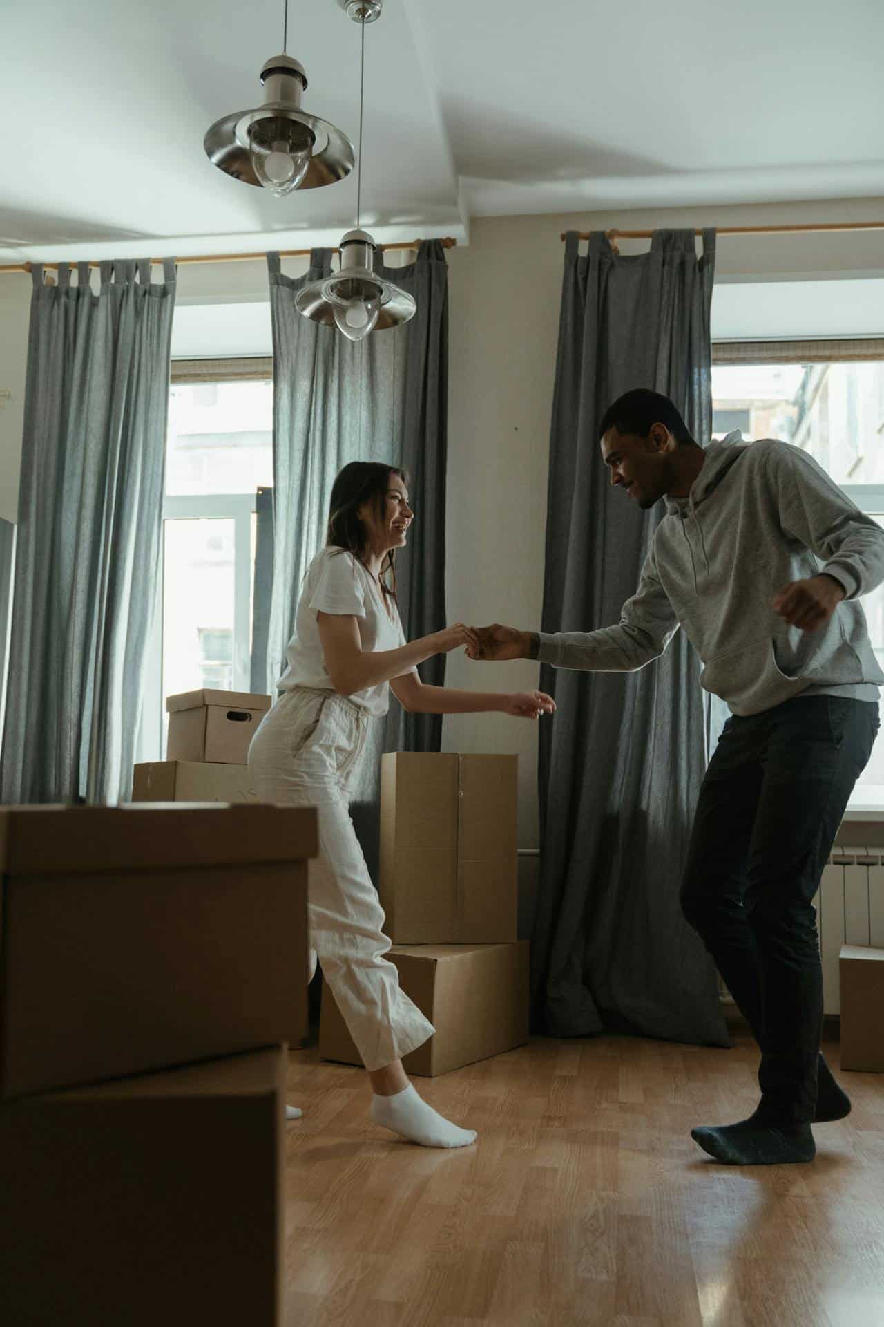 A couple dancing together in their living room. They are dressed in sweats and surrounded by boxes, as though they are in the process of moving into a new apartment. 
