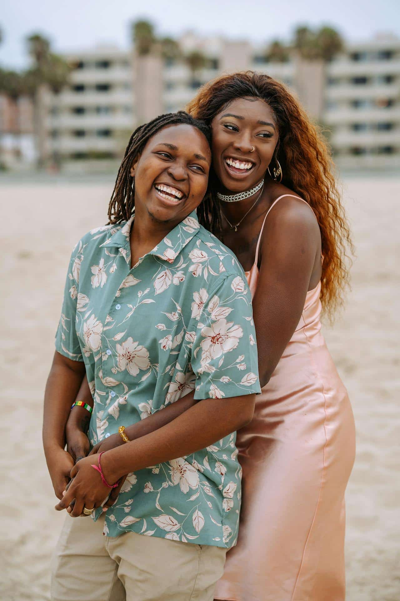 A Black lesbian couple poses together, looking happy. One is wearing a pink dress, the other is wearing a blue button up shirt with floral print. The beach can be seen blurred in the background.