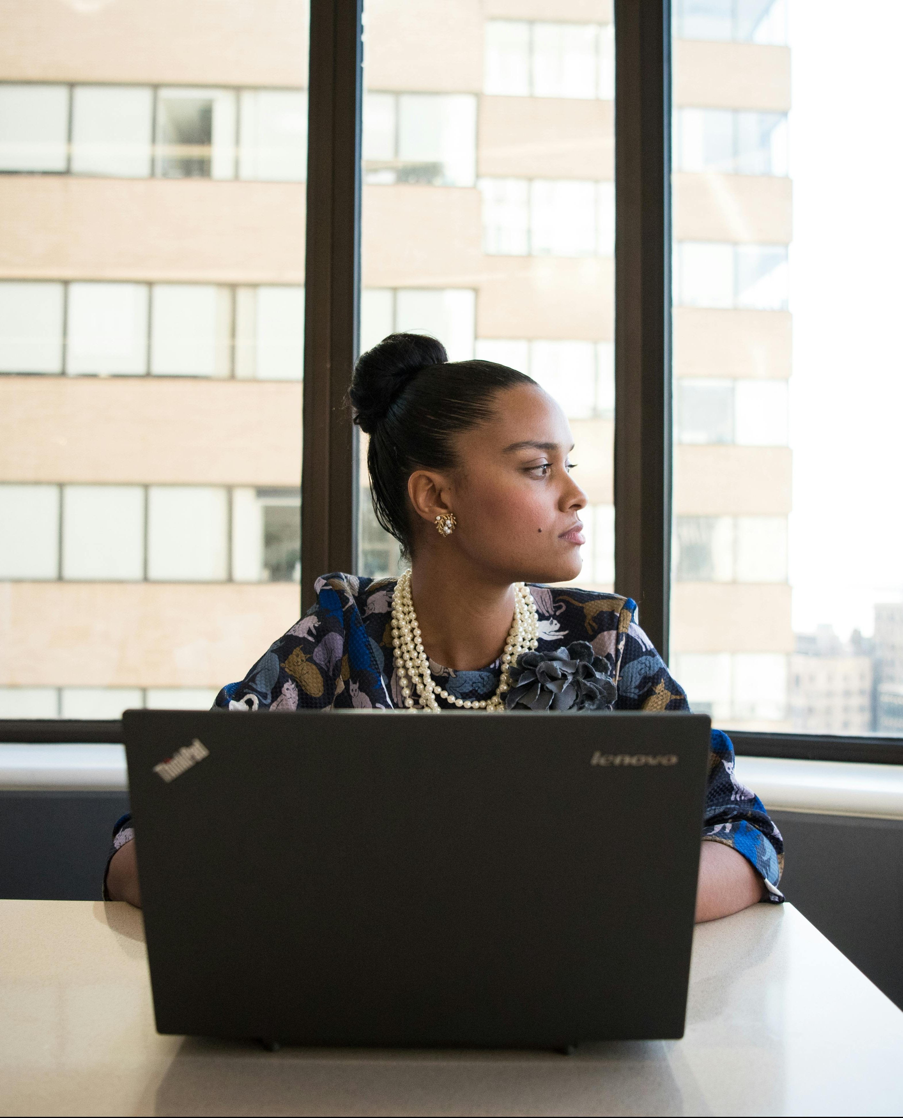 a young Black woman sitting at her laptop, staring out of the window in her office