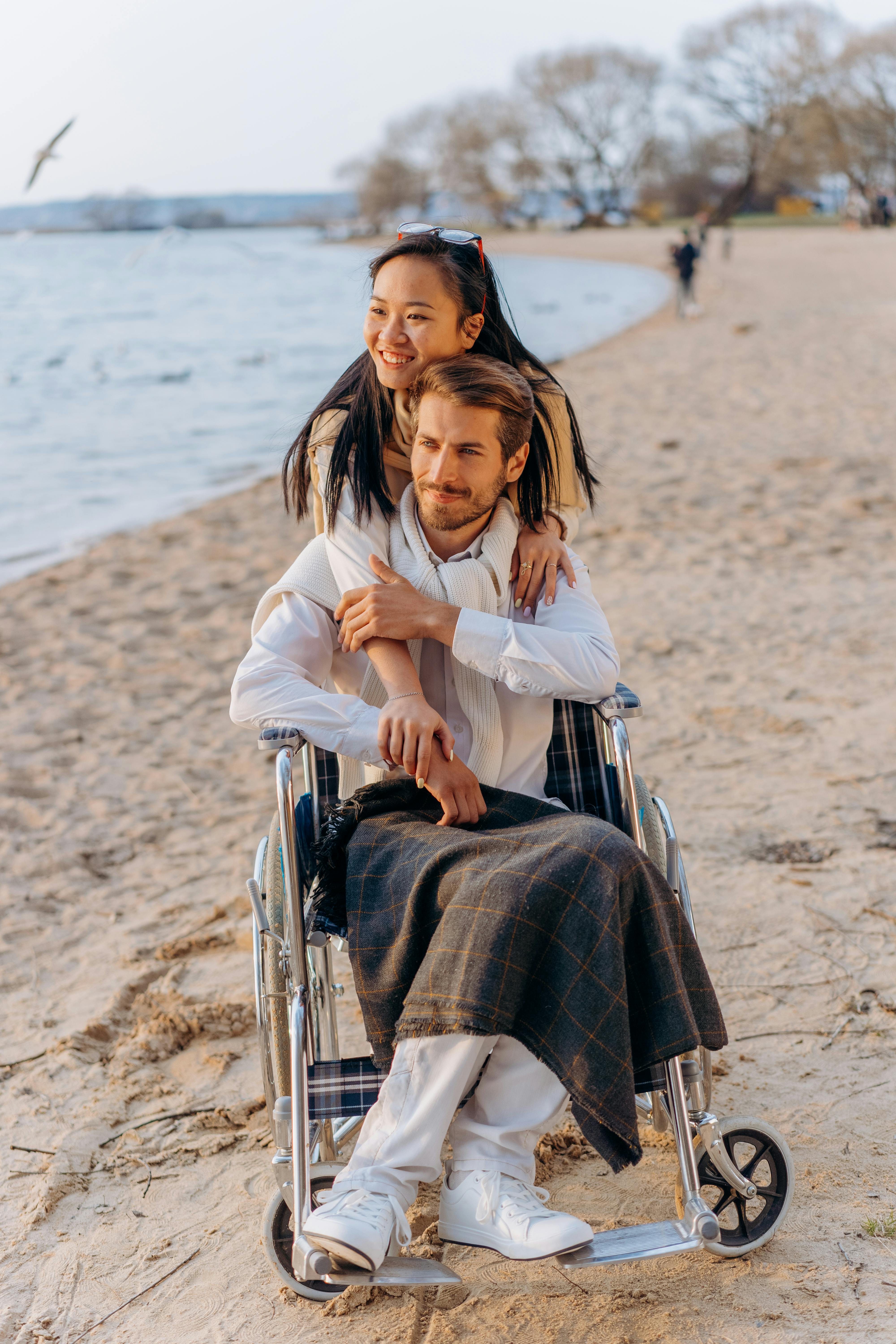 A couple posing on the beach. One partner, a white male, sits in a wheelchair while other partner, an Asian woman, stands hugging him from behind. 