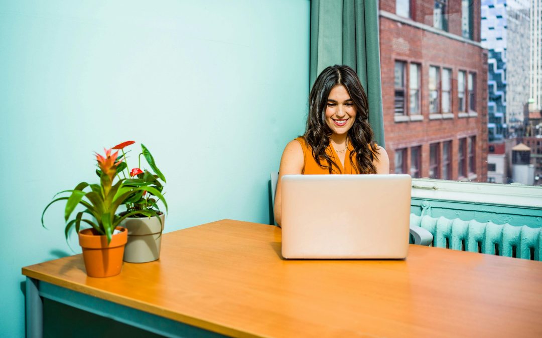 A Latina woman with long wavy dark hair, sitting in a light teal room at a table in front of a window showing a street, working at a laptop while smiling.