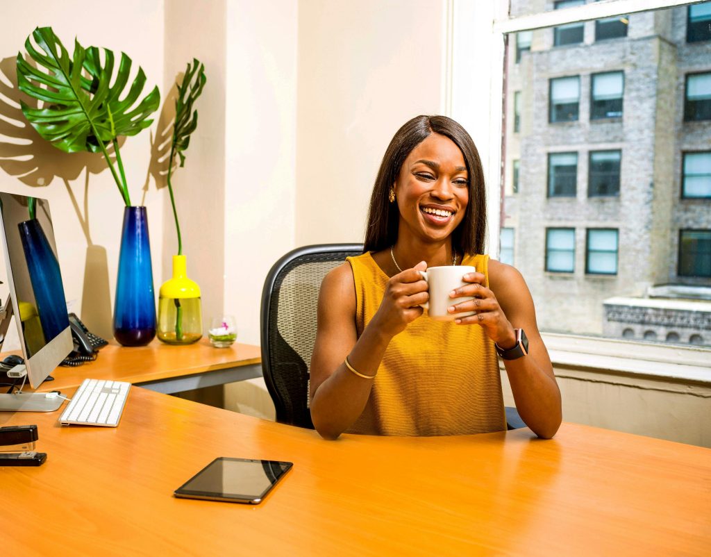 A Black woman with shoulder length hair sitting at a desk in an office, holding a cup of coffee and smiling.