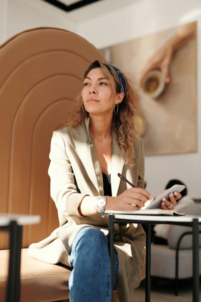 A woman with medium length wavy hair sitting in a cafe, writing in a notepad.