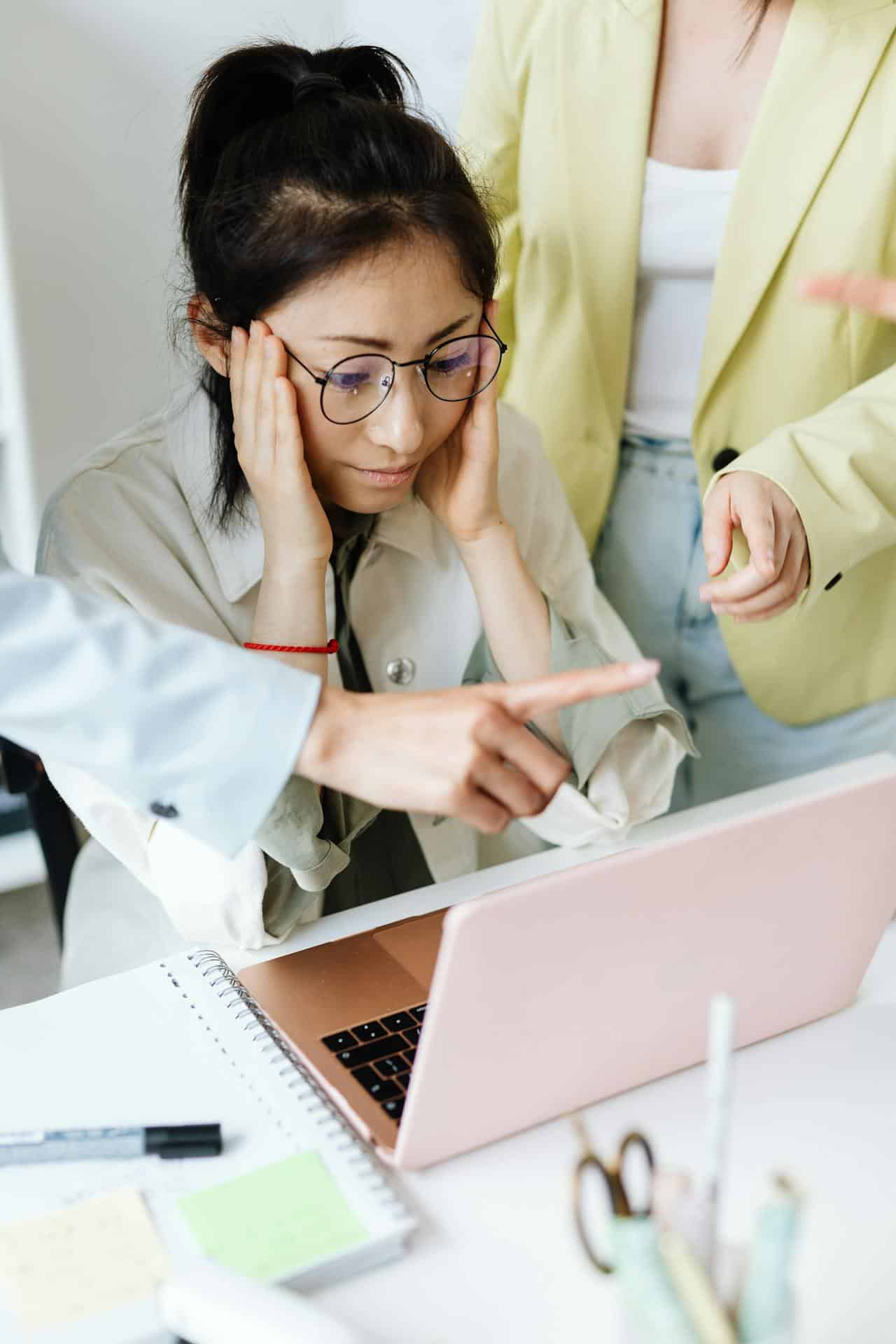 A young Asian woman sits at her laptop with stressed or anxious feelings, while two coworkers stand near her pointing at her laptop, as though criticizing or micromanaging her. 