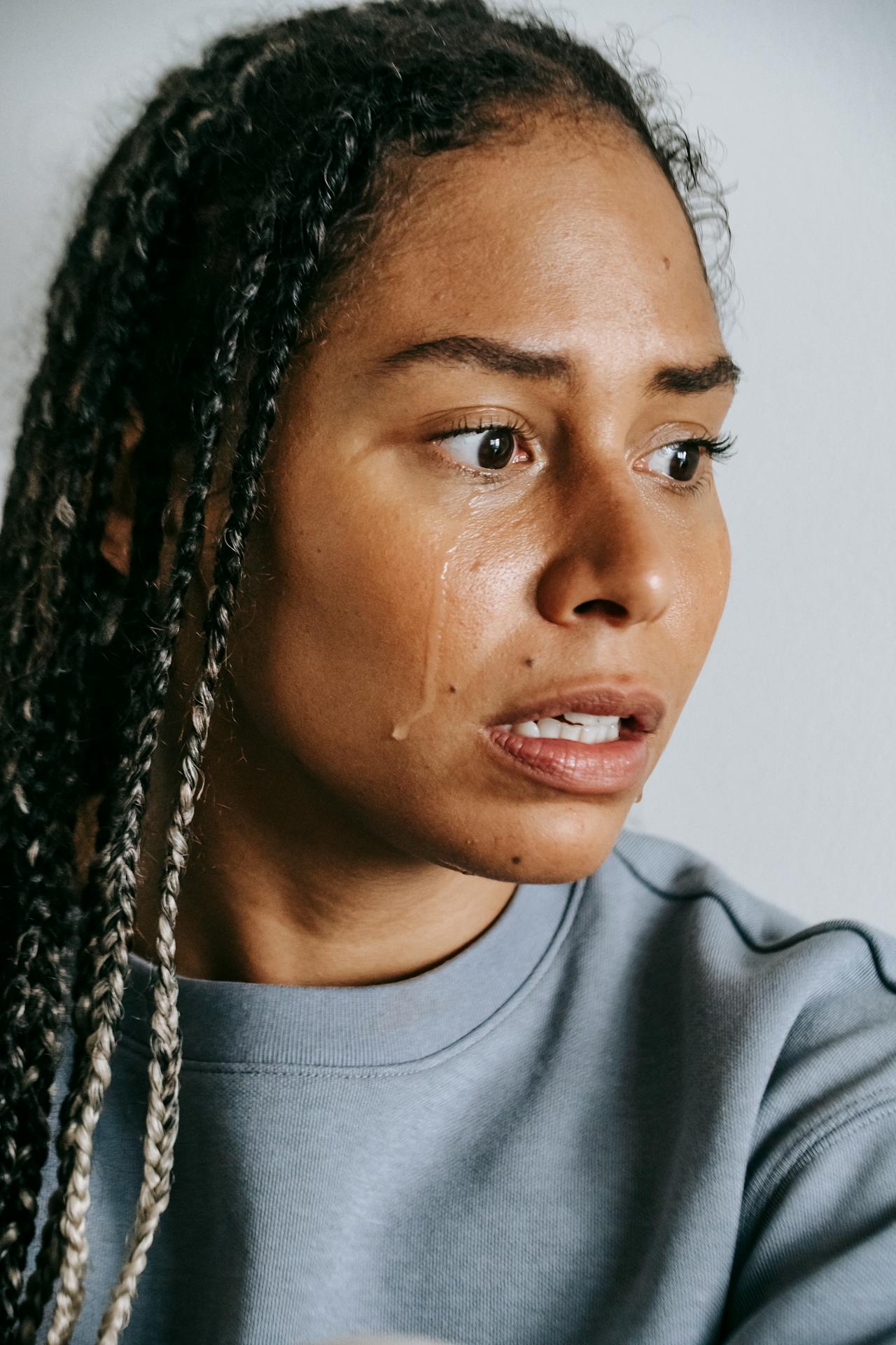 Stock photo of Black woman with long braids, photographed from shoulders up. She is wearing a gray sweatshirt and looking away from the camera, tears running down her face.