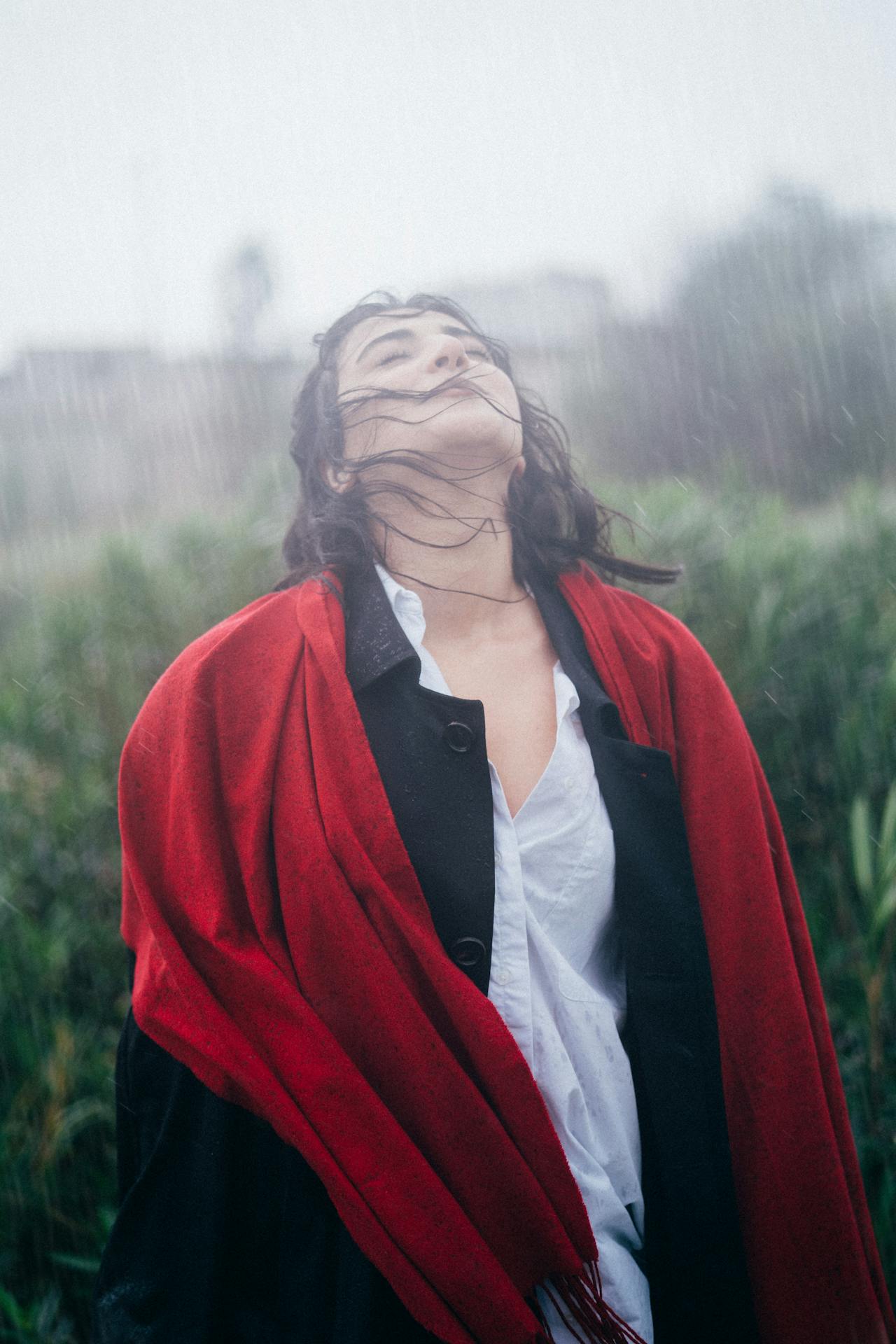 A light skinned woman with medium brown hair stands in the rain, wearing a red coat. Her face is turned upwards and her eyes are closed.