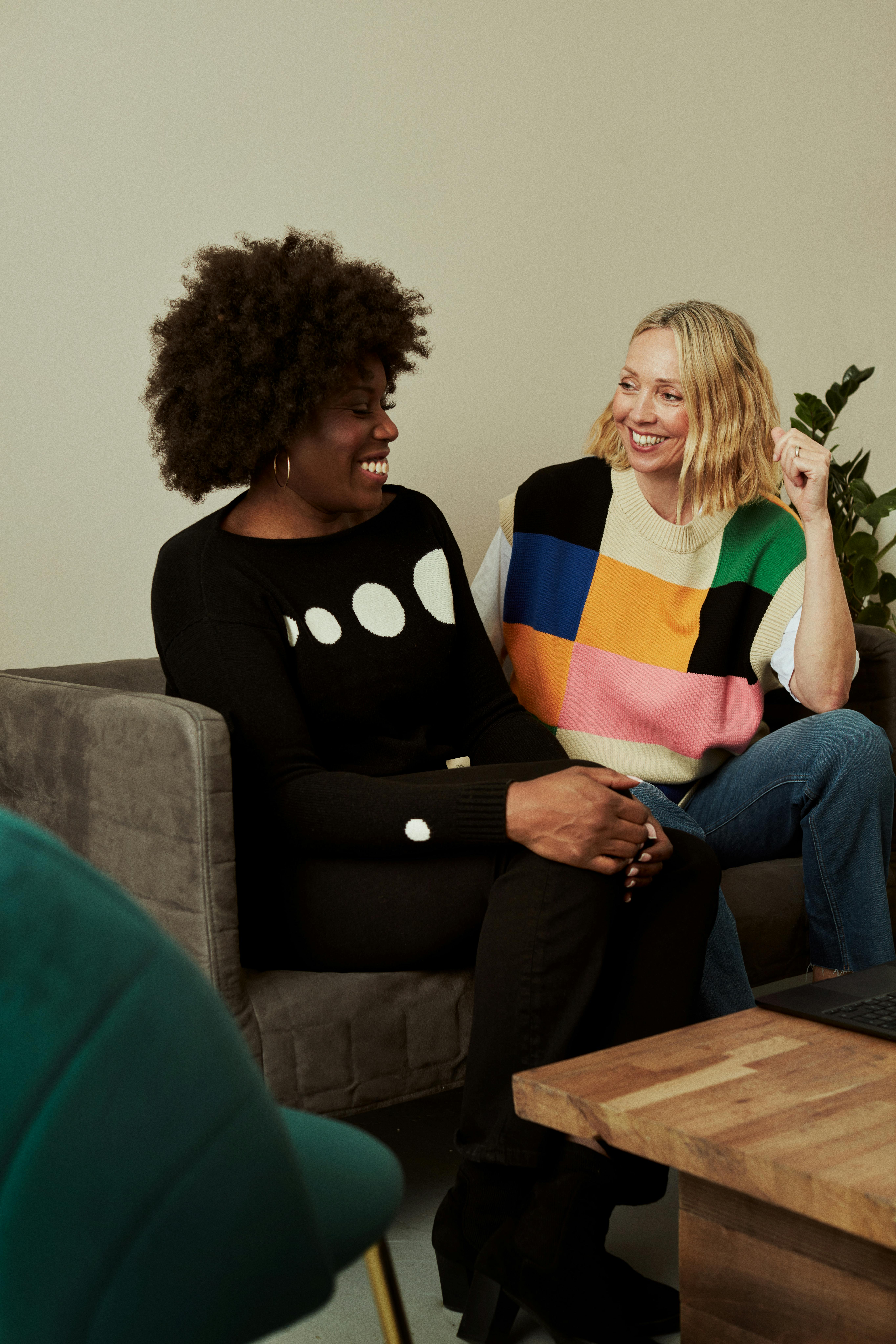 A Black woman and a white woman smiling at one another, sitting on a couch in a therapists's office. 
