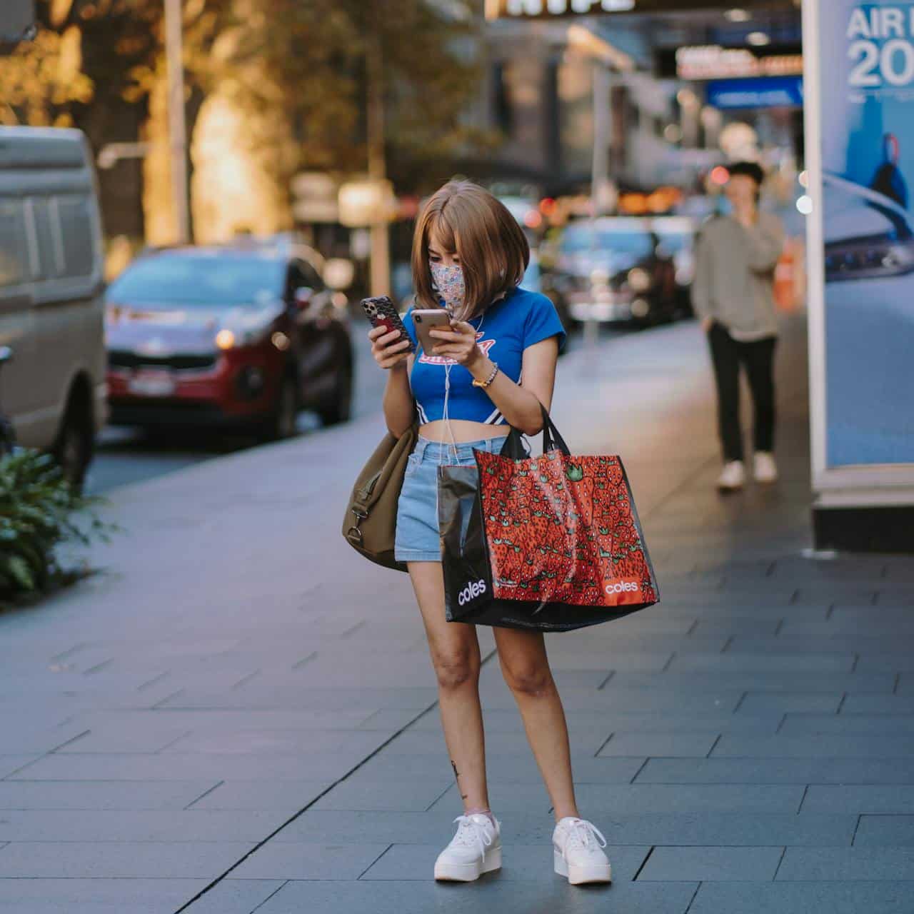 A young woman standing on a sidewalk looking at her phone, with shopping bags on her arms. She is wearing a mask to protect herself from Covid.