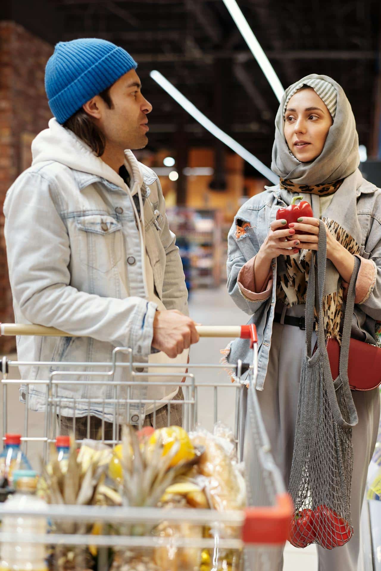 A couple shopping together at a grocery store. A man pushes a partially full grocery cart while a woman in a hijab holds a red bell pepper. They appear to be in conversation.