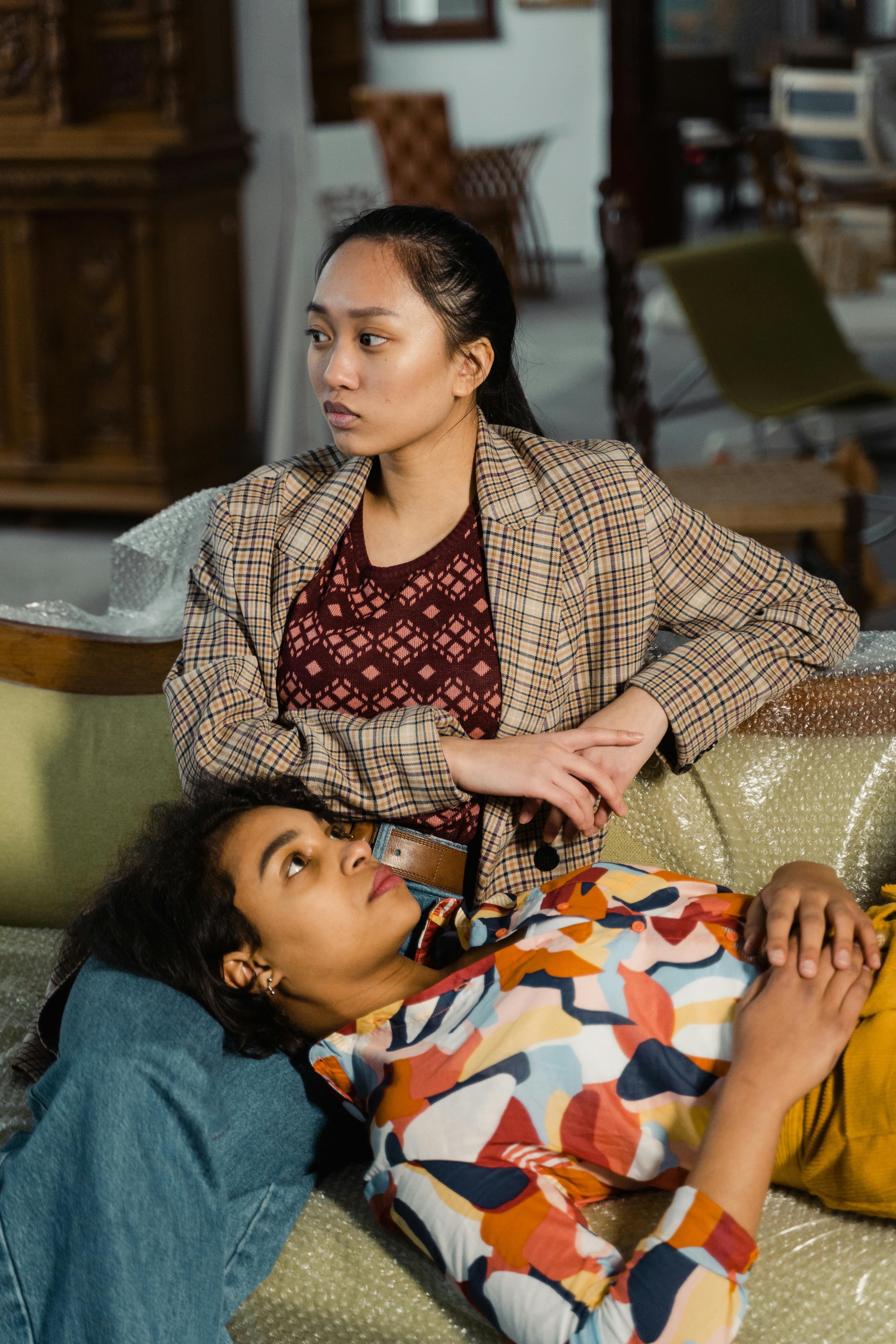 Two women of color sitting on a couch; one has her head resting on the other's lap. 