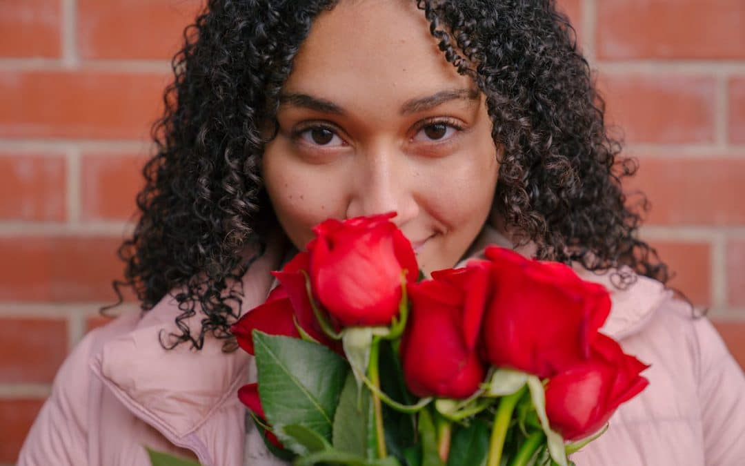 A Black woman with curly long hair stares at the camera while smelling red roses.