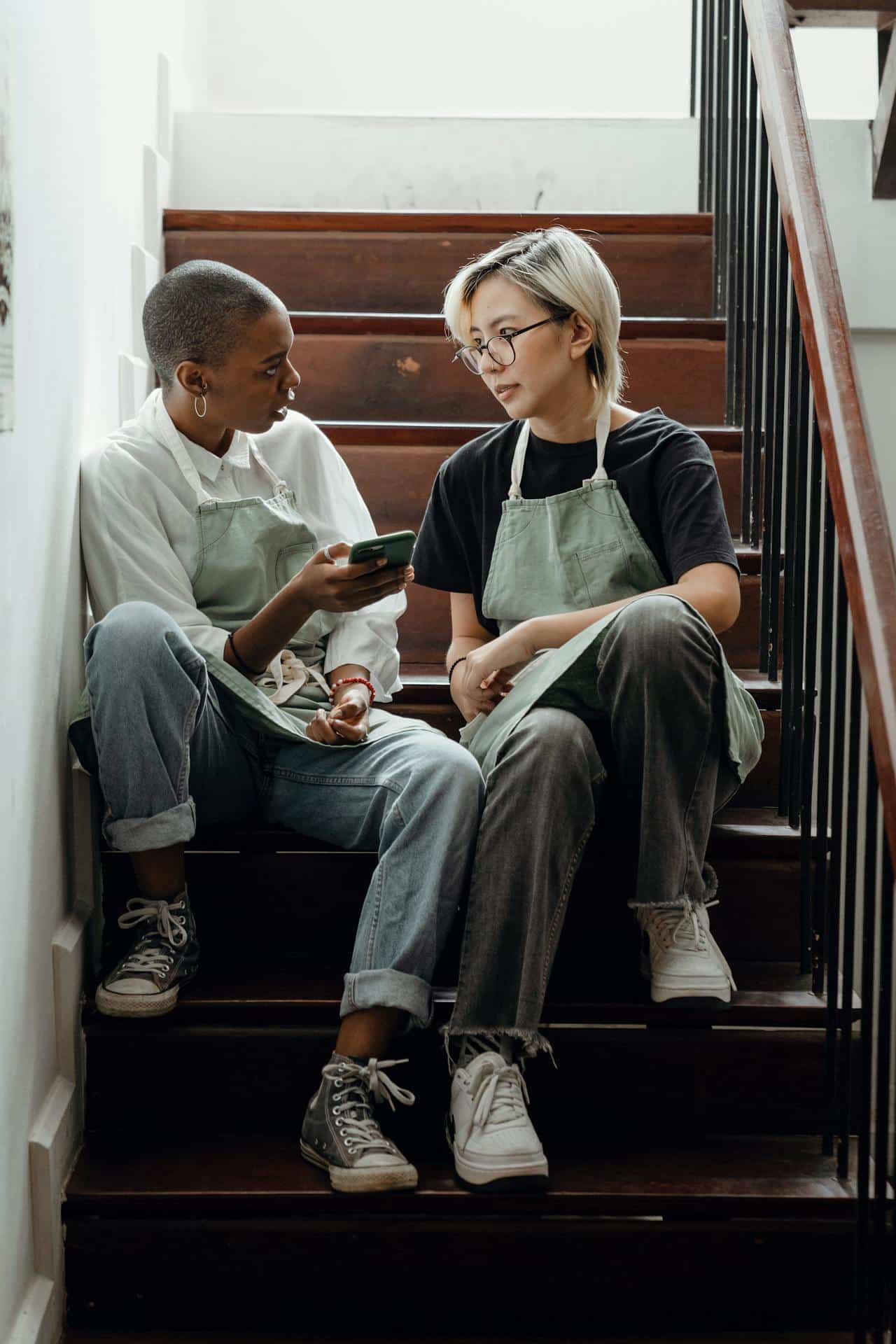 A black woman with a shaved hairstyle holding a cellphone and an Asian woman with bleached short hair sit together on a stairwell, talking. 