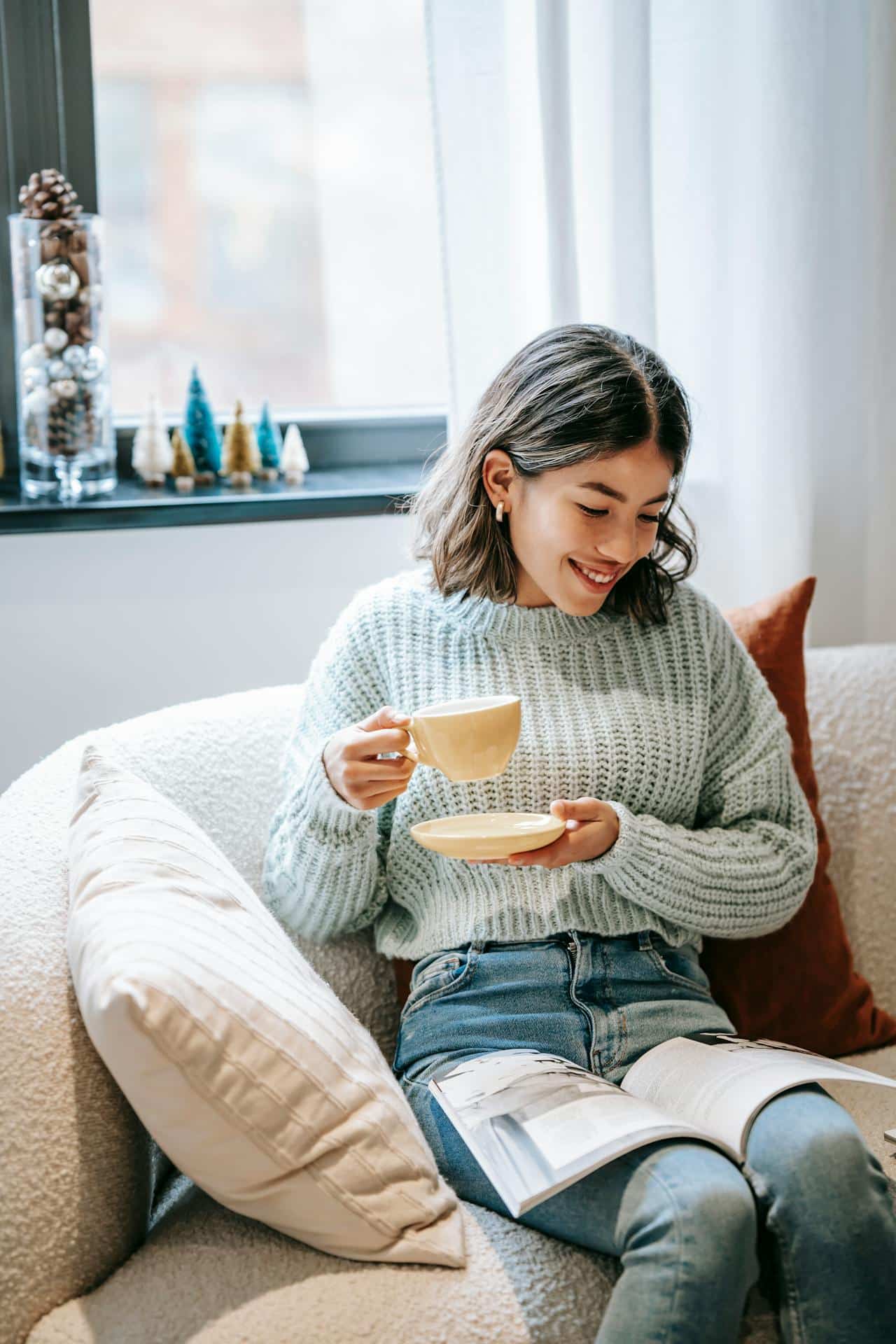 A Latina woman sits on a couch reading a magazine while drinking coffee.