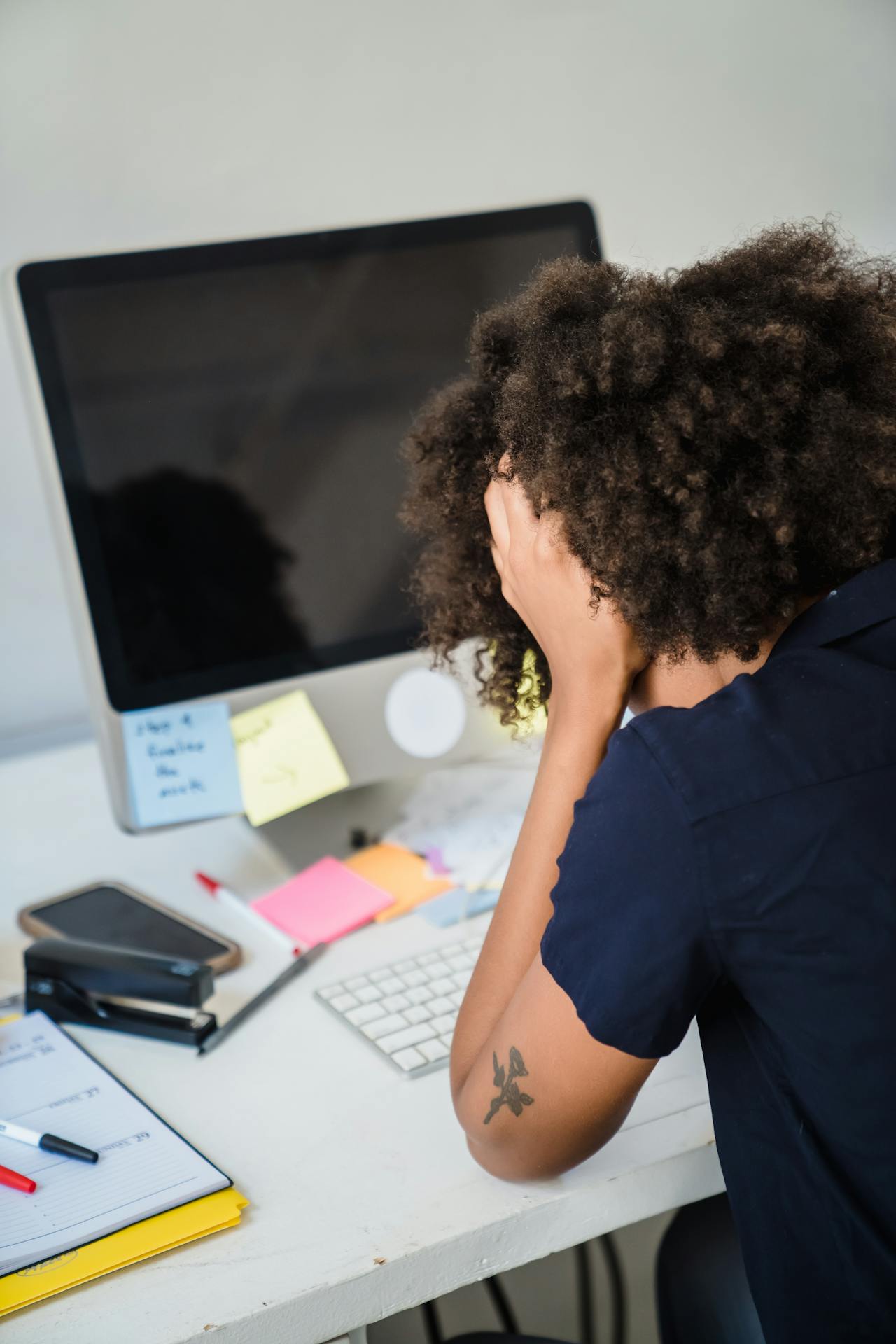 Black woman with afro hairstyle sits with her head in her hands in front of her work desk. 