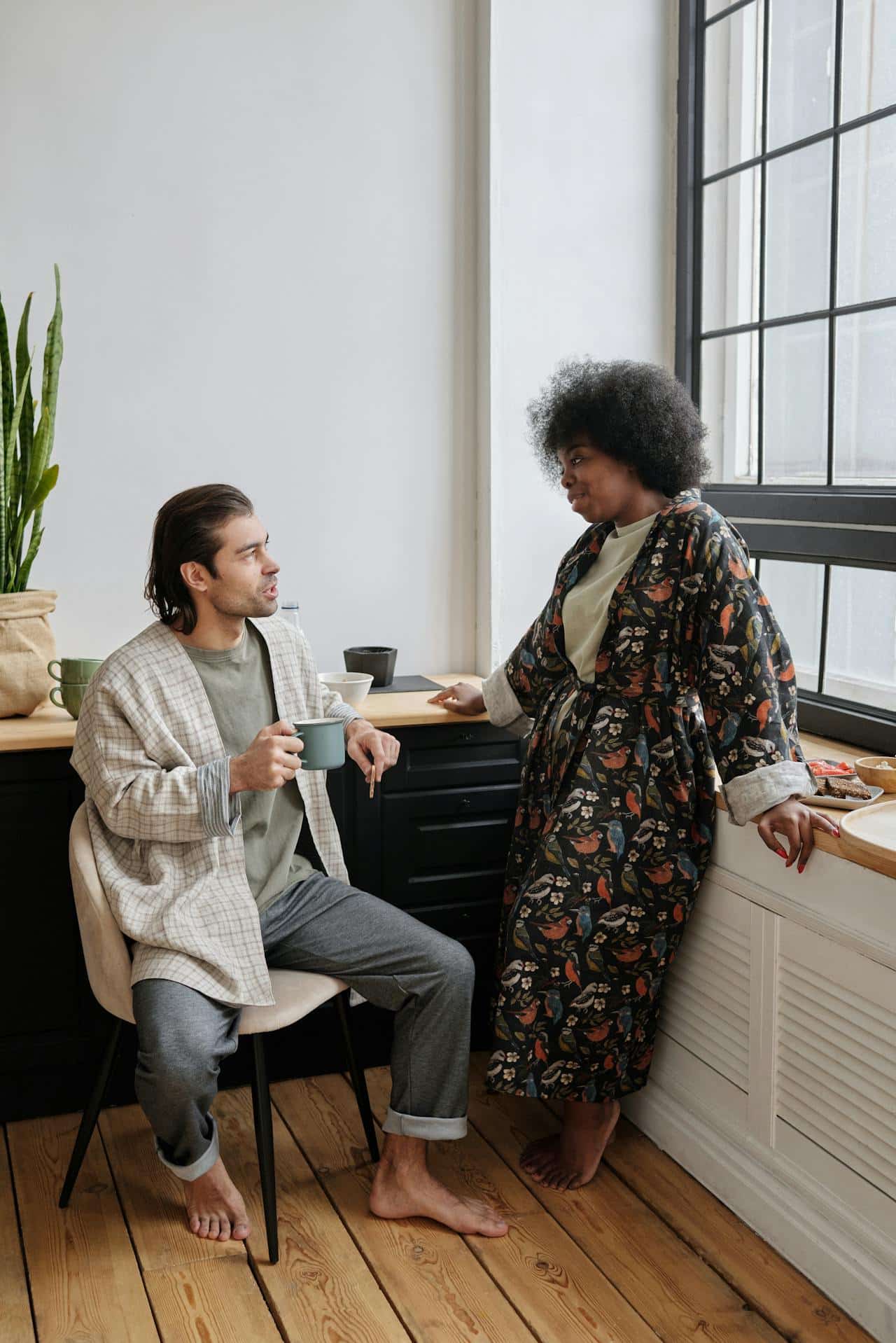 A couple in their home, talking. A white presenting man with dark hair in casual attire sits, while speaking with a Black woman wearing a bathrobe, standing by the window.