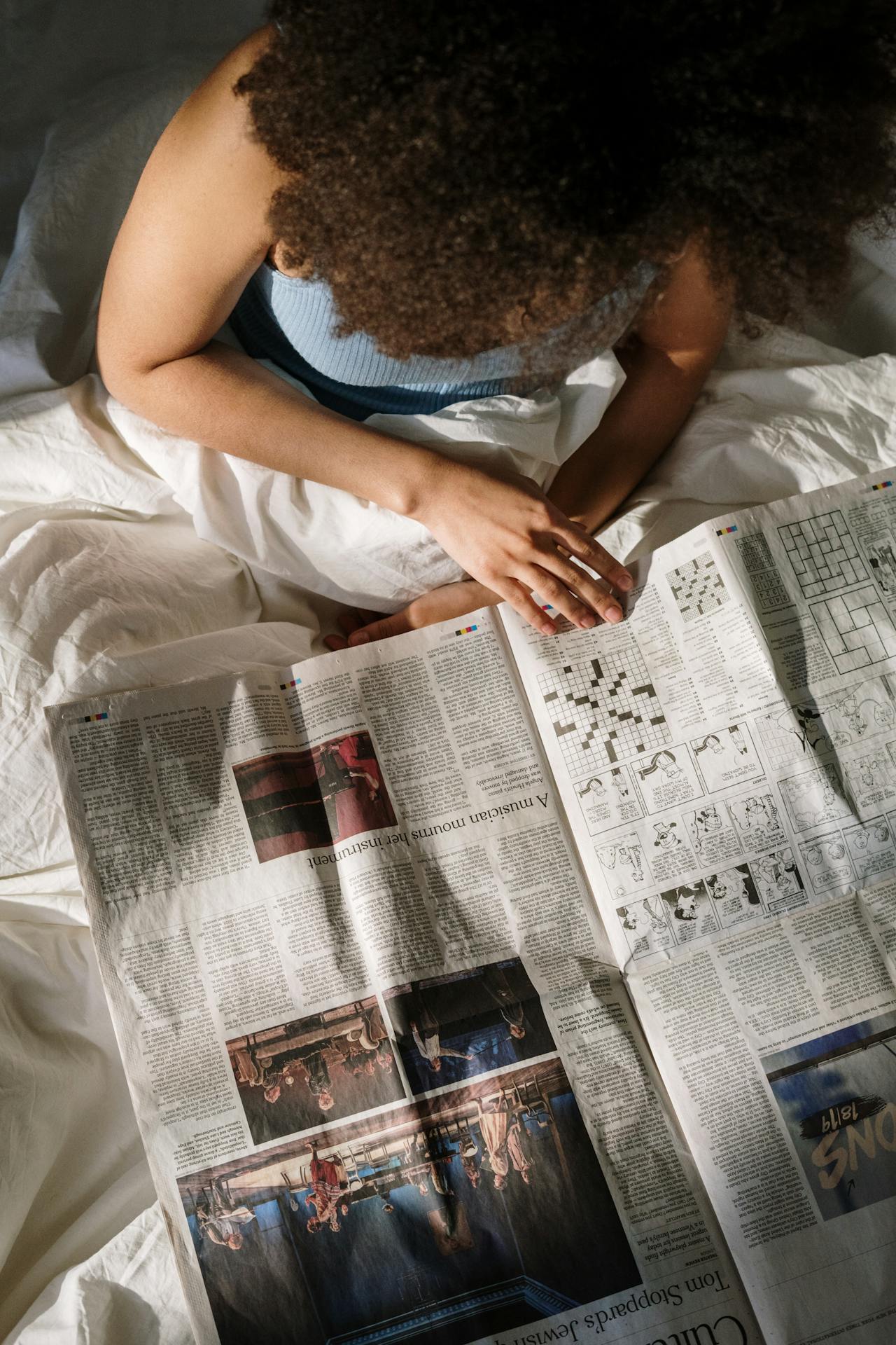 An overhead view of a black woman with an afro reading a newspaper in bed. 