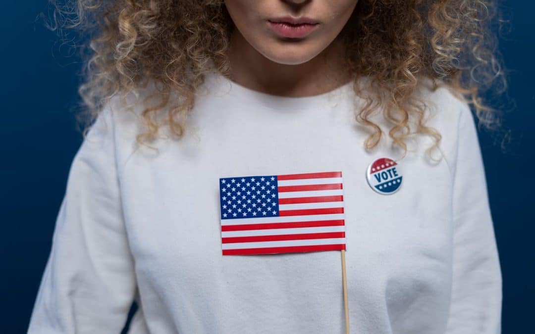 Half of a tan woman's is visible; she has blonde curly hair, wearing a white sweatshirt with a "Vote" button, while holding a small American flag.