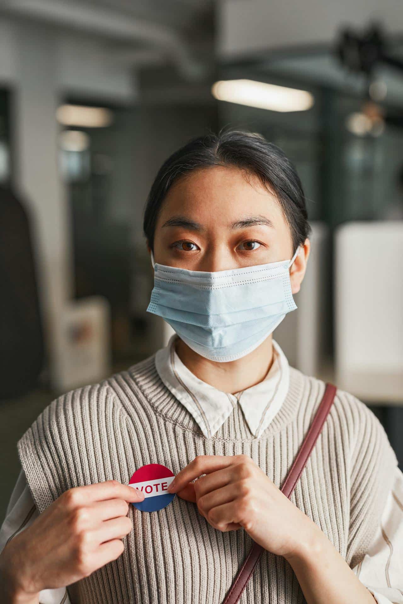 An Asian woman with her hair tied back, wearing a face mask and sweater with a collar, holds a "Vote" sticker on herself. 