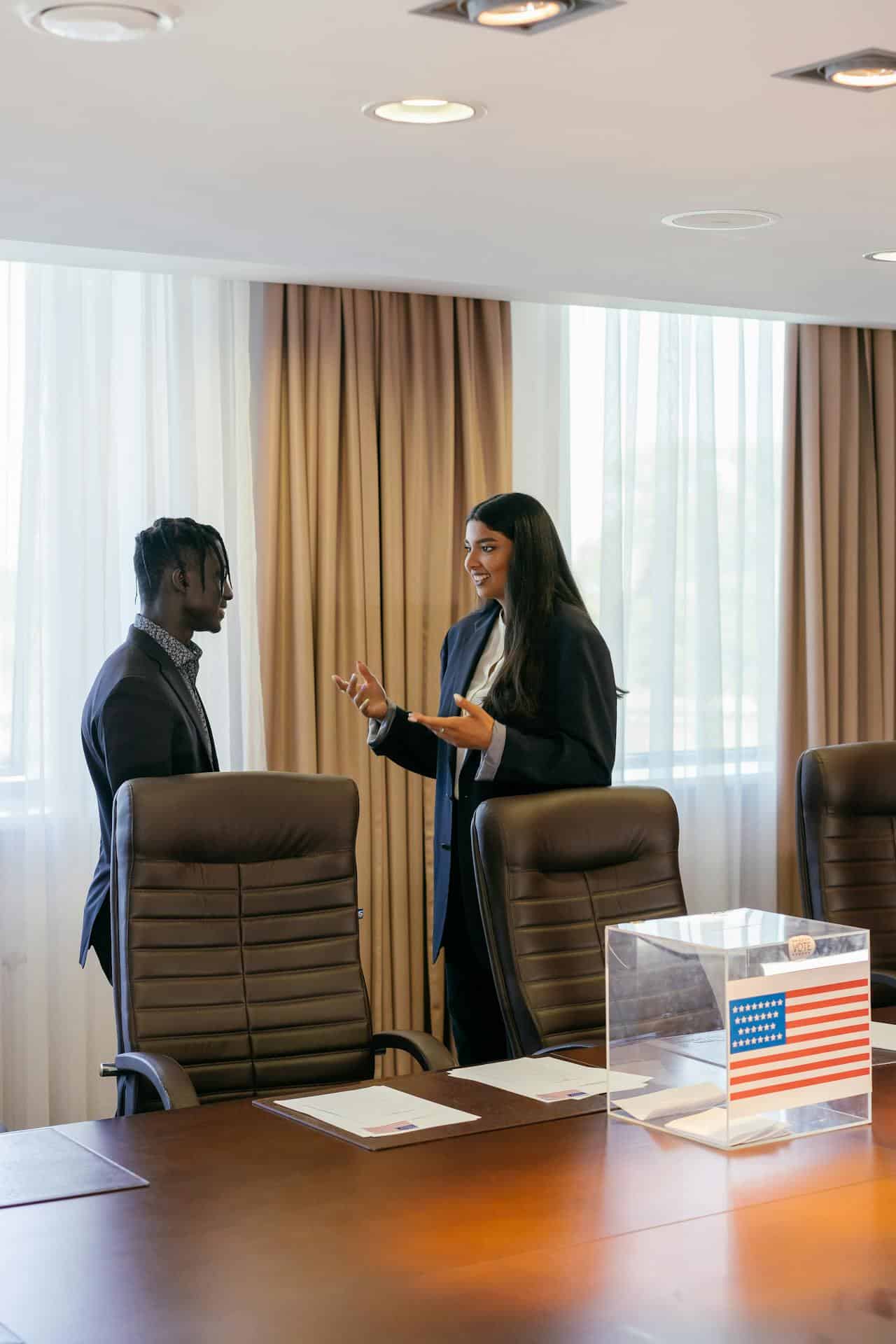 A black man and Indian woman dressed in professional attire smile while in discussion. Tan and sheer curtains are behind them, and brown office chairs, a table with a ballot box are in front of them.