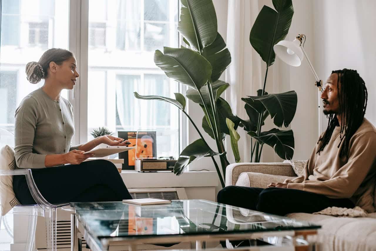 A therapist with curly hair tied back, wearing a light green sweater, sits on a transparent chair holding a clipboard, gesturing as she speaks. The client, a man with long locks, dressed in a beige sweater, listens attentively while seated on a sofa.