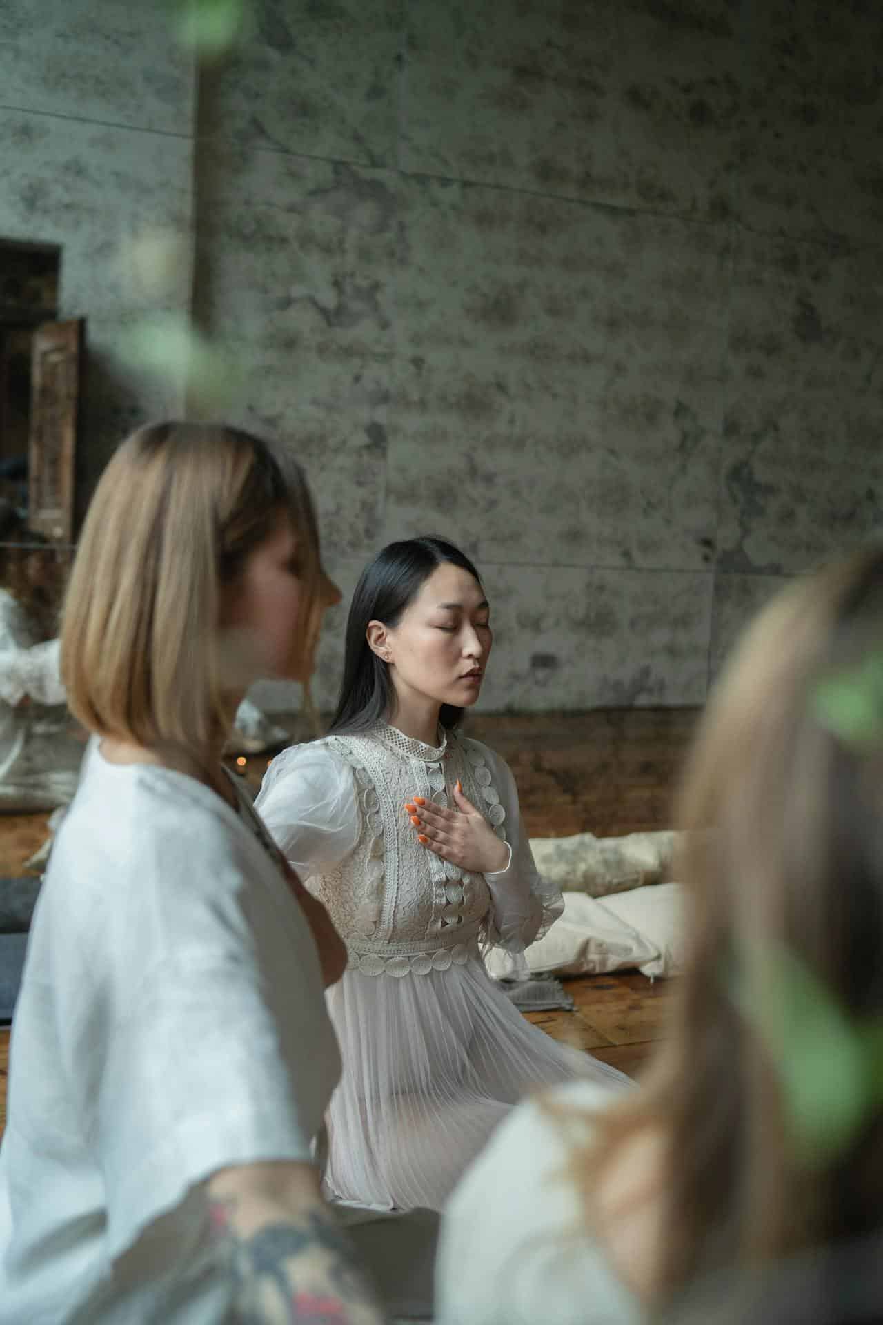 A group of women practicing mindfulness or meditation indoors. A young Asian woman, with straight black hair and orange-painted nails, wears a white lace dress and has her hand on her chest with her eyes closed.