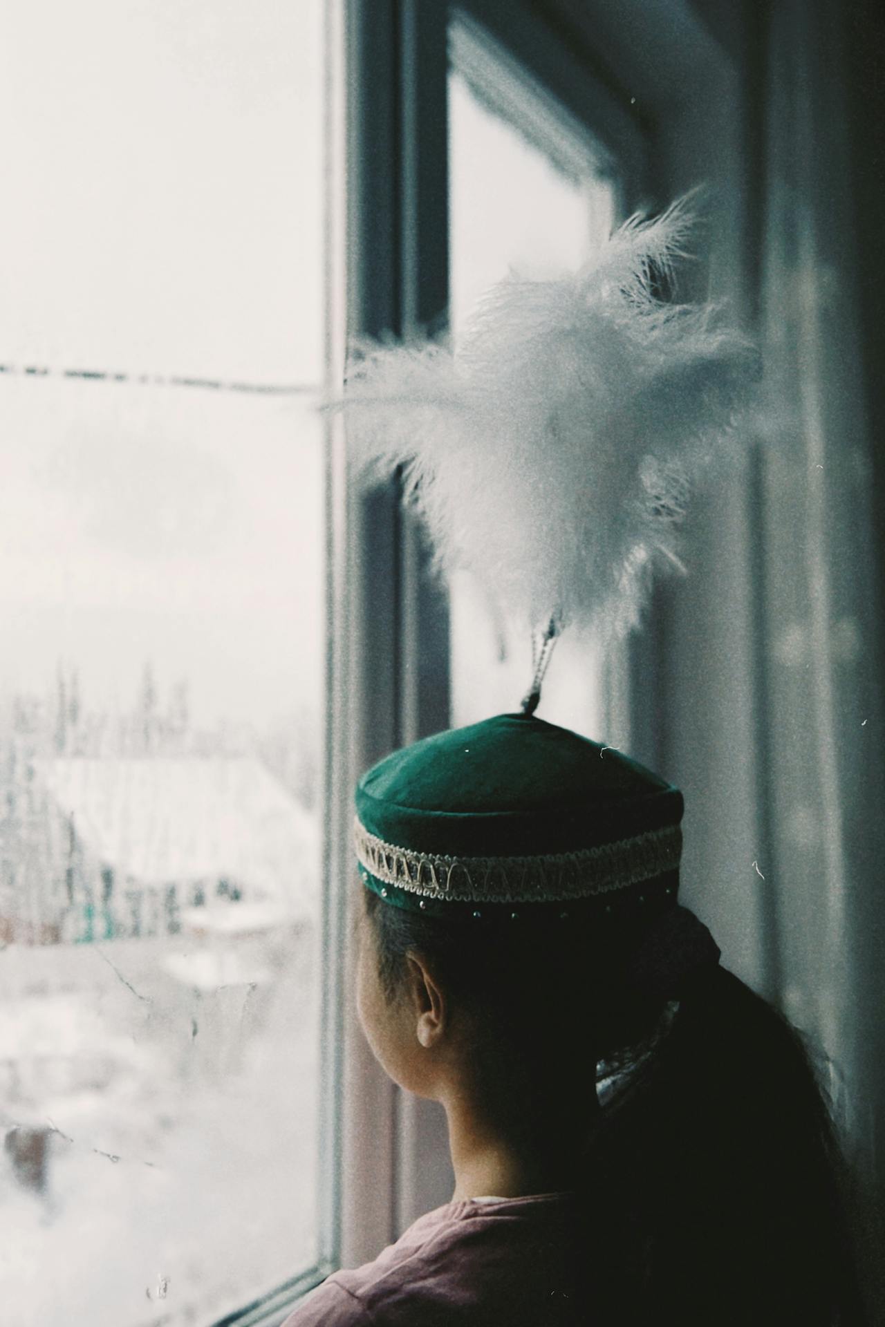  Asian girl wearing traditional green Kazahk hat, staring out the window at a winter scene.