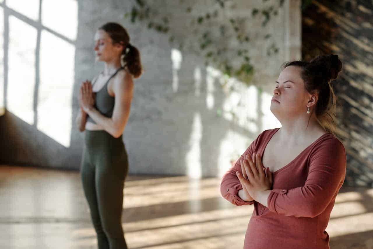 Two Woman stand in a sunlit room doing a standing breathing meditation